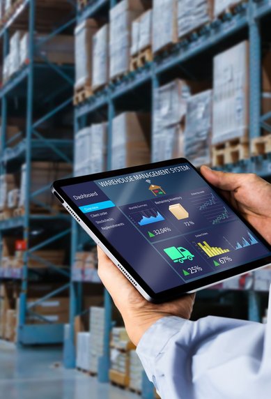 A person is holding a tablet with data analytics in a warehouse, surrounded by shelves of goods.