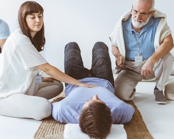 Eine Frau führt eine Behandlung an einem liegenden Mann durch, während ein älterer Mann mit Brille und Notizen daneben steht. Im Hintergrund sind weitere Personen in einer ähnlichen Umgebung zu sehen.