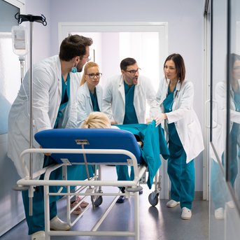 Eine Gruppe von fünf medizinischen Fachkräften in blauen Scrubs und weißen Kitteln, die einen Patienten auf einer Trage in einem Krankenhauszimmer betreuen.