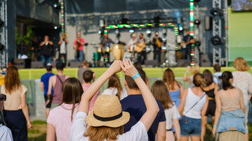 Eine Menschenmenge schaut auf eine Bühne, auf der eine Band auftritt. Eine Person mit einem Strohhut hebt die Hände in die Luft.