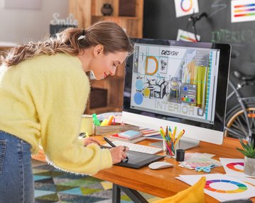 Eine Frau in einem gelben Pullover arbeitet an einem Grafiktablett, während sie auf einen Computerbildschirm mit Innendesign-Layouts schaut.
