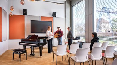 A group of four people is standing in a music room with a piano and a screen. Two of them are holding instruments.