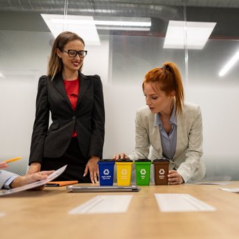 Two women in business attire are standing at a table with coffee cups in various colors. Other people are visible at the table.