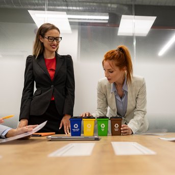 Zwei Frauen in Geschäftskleidung stehen an einem Tisch mit Kaffeetassen in verschiedenen Farben. Weitere Personen sind am Tisch sichtbar.