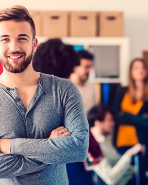 Ein Mann mit Bart lächelt und steht mit verschränkten Armen im Vordergrund, während eine Gruppe von Menschen im Hintergrund miteinander spricht.