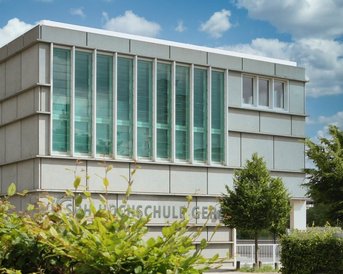 Moderne Schulgebäude mit großen Fenstern und einer klaren Fassade. Im Vordergrund sind grüne Pflanzen und Bäume sichtbar. Der Name der Schule ist an der Wand angebracht.