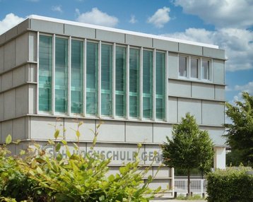 Moderne Schulgebäude mit großen Fenstern und einer klaren Fassade. Im Vordergrund sind grüne Pflanzen und Bäume sichtbar. Der Name der Schule ist an der Wand angebracht.
