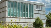 Moderne Schulgebäude mit großen Fenstern und einer klaren Fassade. Im Vordergrund sind grüne Pflanzen und Bäume sichtbar. Der Name der Schule ist an der Wand angebracht.