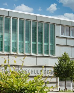 Moderne Schulgebäude mit großen Fenstern und einer klaren Fassade. Im Vordergrund sind grüne Pflanzen und Bäume sichtbar. Der Name der Schule ist an der Wand angebracht.