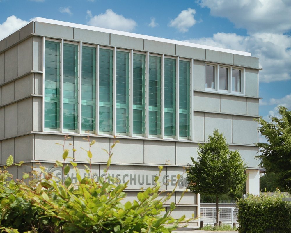 Moderne Schulgebäude mit großen Fenstern und einer klaren Fassade. Im Vordergrund sind grüne Pflanzen und Bäume sichtbar. Der Name der Schule ist an der Wand angebracht.