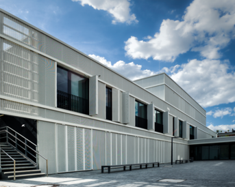 Ein modernes Gebäude mit einer hellen, strukturierten Fassade und großen Fenstern, umgeben von einem klaren Himmel mit einigen Wolken. Eine Treppe führt zum Eingang.