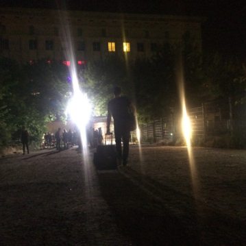 A person with a suitcase walks along a lit path, surrounded by trees and other people, at night.