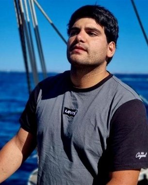 Ein Mann steht auf einem Boot, trägt ein graues T-Shirt mit schwarzem Ärmel und blickt nachdenklich in die Ferne, während das Meer im Hintergrund sichtbar ist.
