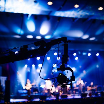Camera crane in the foreground, with a band performing on a lit stage. Colorful lights and a festive atmosphere.