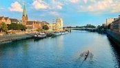 Blick auf einen Fluss mit einem Ruderboot, umgeben von historischen Gebäuden und einer Kirche im Hintergrund.