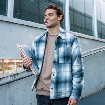 Young man in a checkered shirt standing on a staircase, holding documents in his hand while smiling.