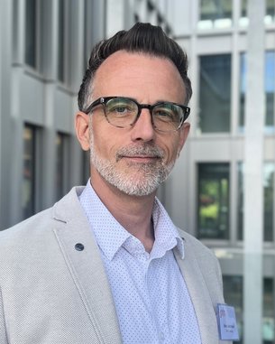 Ein Mann mit Brille und grauem Bart trägt einen hellen Anzug und ein gepunktetes Hemd. Er steht vor einem modernen Gebäude und schaut direkt in die Kamera.