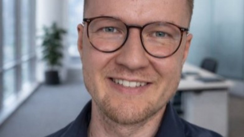 Ein Mann mit Brille lächelt in einem modernen Büro. Im Hintergrund sind Fenster und eine Pflanze sichtbar.