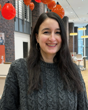 Eine junge Frau mit langen, dunklen Haaren trägt einen grauen Pullover und lächelt. Im Hintergrund hängen rote Lampen.