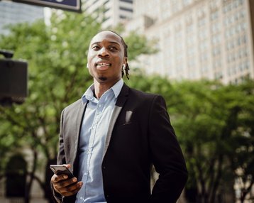 Kategorie New Work Studium studieren SRH University Ein Mann in einem Anzug steht an einer Straßenecke mit einem Smartphone in der Hand. Im Hintergrund ist ein Straßenschild mit der Aufschrift "Fifth Ave" und grüne Bäume zu sehen.