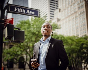 Kategorie New Work Studium studieren SRH University Ein Mann in einem Anzug steht an einer Straßenecke mit einem Smartphone in der Hand. Im Hintergrund ist ein Straßenschild mit der Aufschrift "Fifth Ave" und grüne Bäume zu sehen.