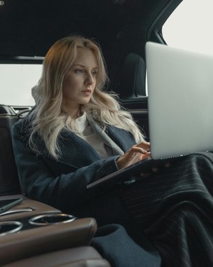 Eine Frau mit langen blonden Haaren sitzt in einem luxuriösen Auto und arbeitet an einem Laptop. Sie trägt einen langen, dunklen Mantel und ist auf den Bildschirm konzentriert. Neben ihr liegt ein Smartphone.