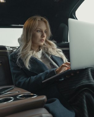 Eine Frau mit langen blonden Haaren sitzt in einem luxuriösen Auto und arbeitet an einem Laptop. Sie trägt einen langen, dunklen Mantel und ist auf den Bildschirm konzentriert. Neben ihr liegt ein Smartphone.