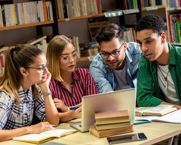 Vier Studierende sitzen an einem Tisch, schauen auf einen Laptop und diskutieren. Bücher liegen offen daneben.