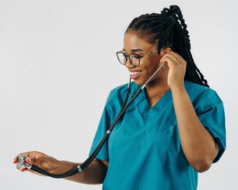 A smiling woman in a blue lab coat holds a stethoscope and listens attentively. She wears glasses and has braided hair. The background is neutral and bright.