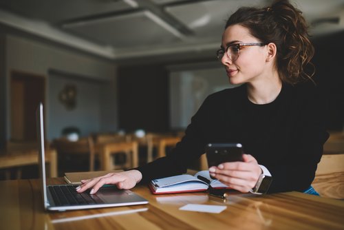 Eine Frau sitzt an einem Tisch, arbeitet am Laptop und hält ein Smartphone in der Hand. Notizbuch und Stift liegen neben ihr.