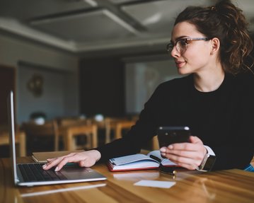 Eine Frau sitzt an einem Tisch, arbeitet am Laptop und hält ein Smartphone in der Hand. Notizbuch und Stift liegen neben ihr.