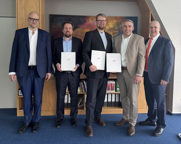 Fünf Männer stehen nebeneinander in einem Büro, halten Dokumente in der Hand und tragen formelle Kleidung. Ein farbiges Gemälde ist im Hintergrund sichtbar.