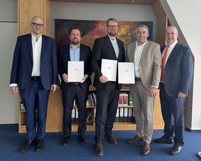 Fünf Männer stehen nebeneinander in einem Büro, halten Dokumente in der Hand und tragen formelle Kleidung. Ein farbiges Gemälde ist im Hintergrund sichtbar.
