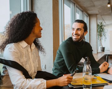 Zwei Personen sitzen an einem Tisch in einem modernen Raum. Eine Frau mit lockigem Haar und ein Mann mit Bart lächeln sich an, während sie Notizen und Schreibutensilien vor sich haben.