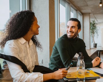 Two people are sitting at a table in a modern room. A woman with curly hair and a man with a beard are smiling at each other, with notes and writing utensils in front of them.
