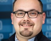 Ein Mann mit Brille und kurzem Bart lächelt in die Kamera, im Hintergrund sind unscharfe Schriftzüge zu sehen.