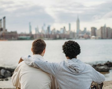 Zwei Personen sitzen am Ufer und blicken auf die Skyline einer Stadt mit Wolkenkratzern im Hintergrund.