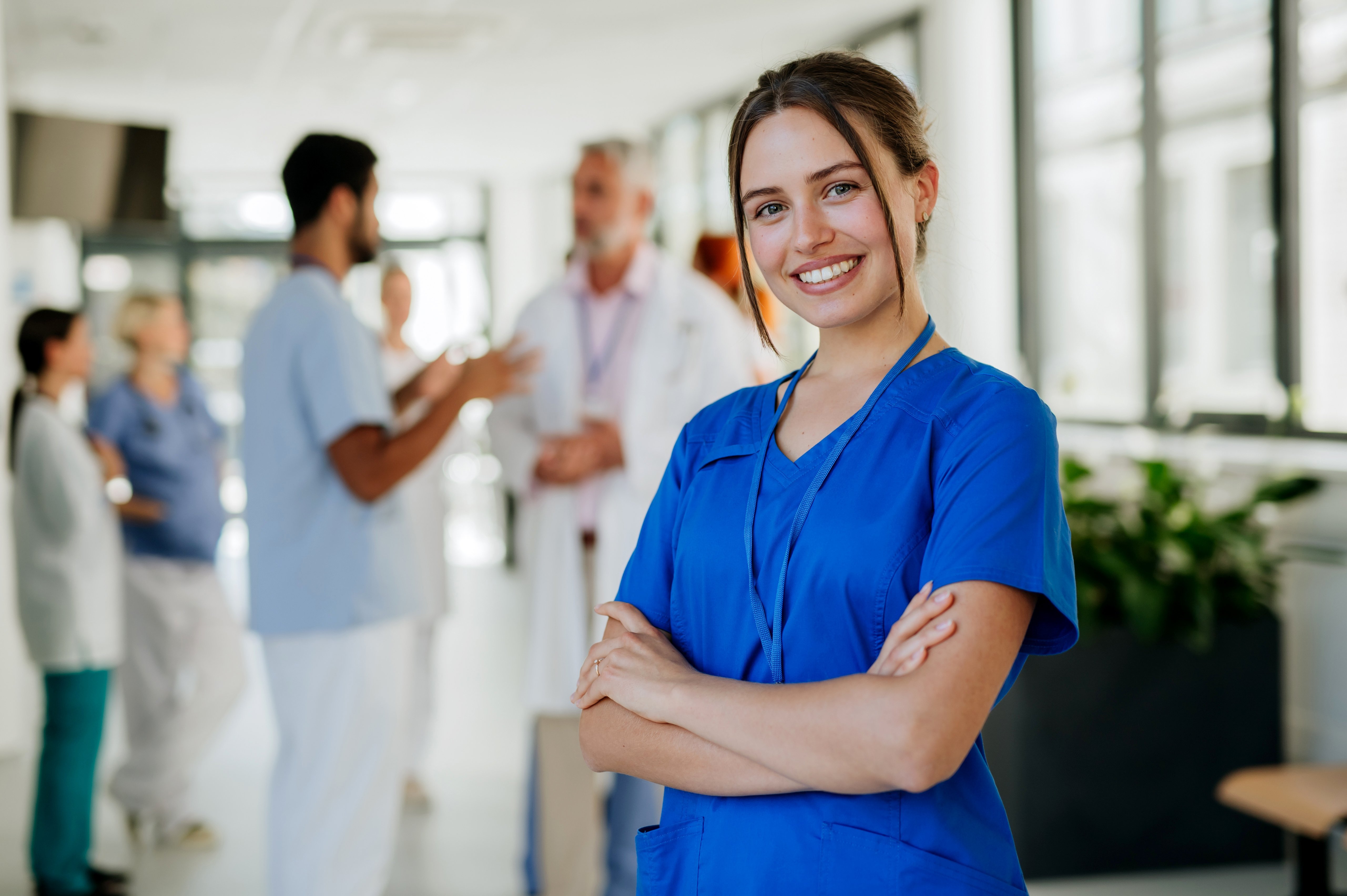 A young woman in blue work attire smiles and stands with her arms crossed in the foreground. In the background, several medical professionals can be seen engaged in conversations, creating a professional atmosphere.