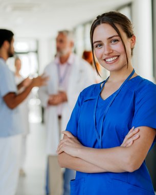 Eine junge Frau in blauer Arbeitskleidung lächelt und steht mit verschränkten Armen im Vordergrund. Im Hintergrund sind mehrere medizinische Fachkräfte in Gesprächen zu sehen, die eine professionelle Umgebung darstellen.