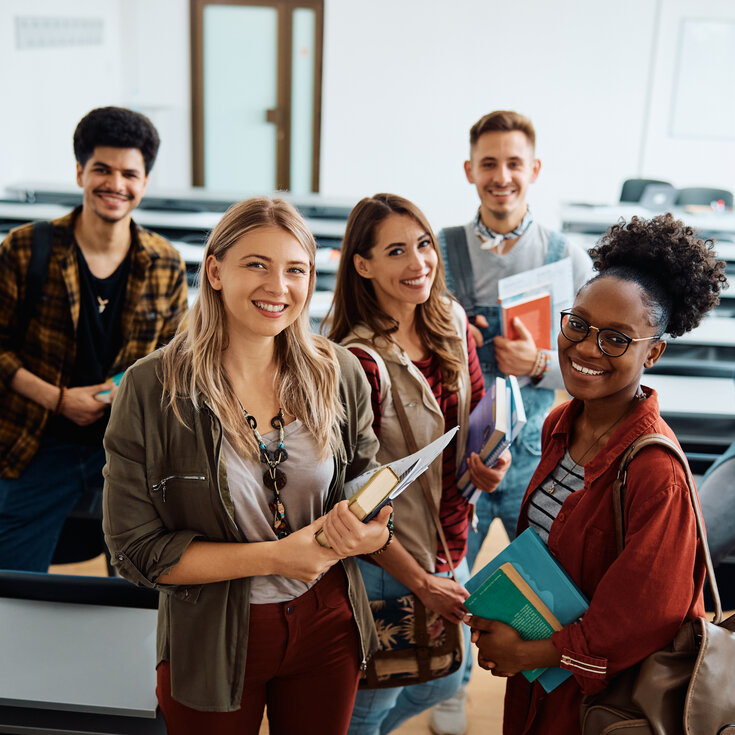 Six students are standing in a classroom, smiling and holding books and notes in their hands.