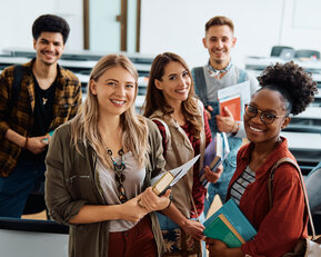 Six students are standing in a classroom, smiling and holding books and notes in their hands.