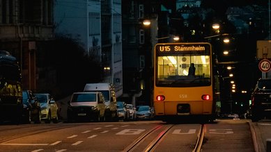 A tram on line U15 heading to Stammheim travels down a well-lit street, surrounded by parked cars and buildings, as dusk begins to set in.