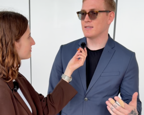 A woman is interviewing a man in a suit as she attaches a microphone to his collar. They are looking at each other.