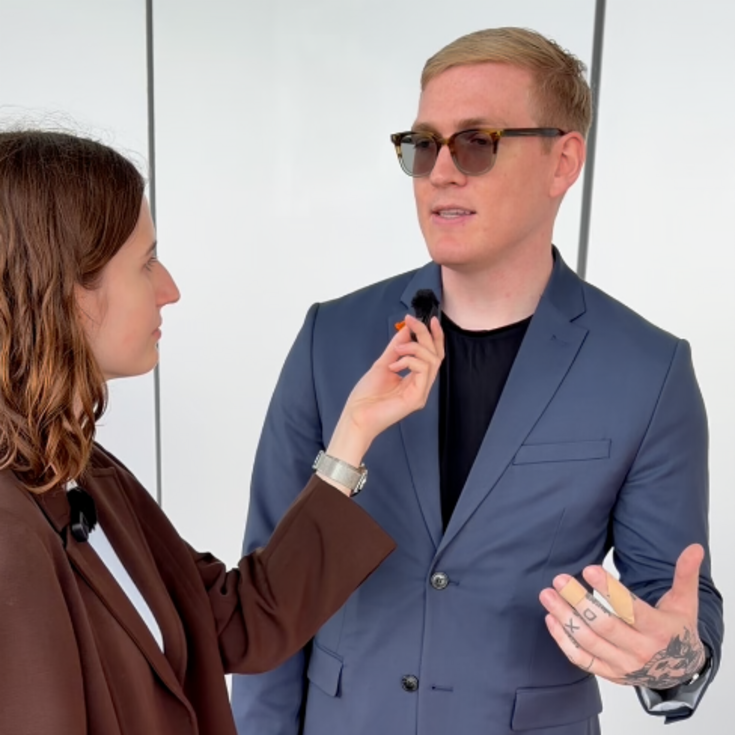 A woman is interviewing a man in a suit as she attaches a microphone to his collar. They are looking at each other.