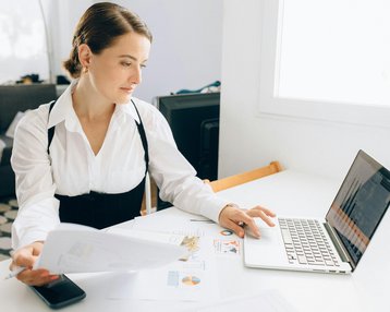 Eine Frau in formeller Kleidung arbeitet an einem Laptop, während sie Dokumente auf einem Tisch durchgeht.