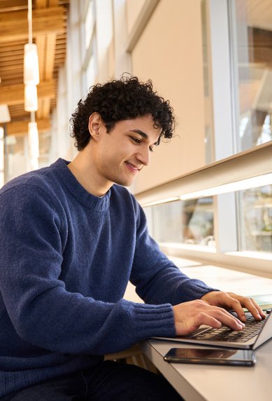 A young man sits at a table in a library, focused on his laptop. He is wearing a blue sweater and smiling as he types. In the background, bookshelves are visible.