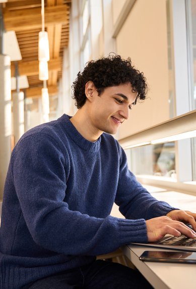 A young man sits at a table in a library, focused on his laptop. He is wearing a blue sweater and smiling as he types. In the background, bookshelves are visible.