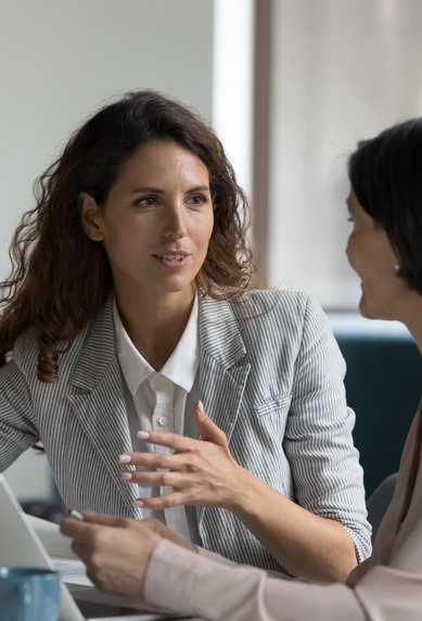 Zwei Frauen führen ein Gespräch am Tisch, eine zeigt auf einen Laptop, während die andere aufmerksam zuhört.