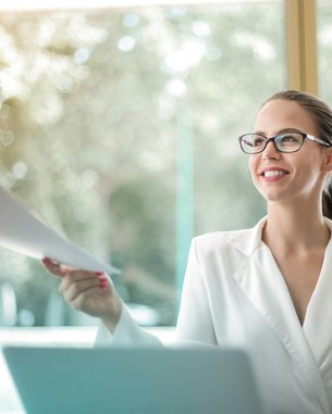 Eine Frau in einem weißen Blazer sitzt an einem Schreibtisch und lächelt, während sie ein Dokument von einer anderen Person entgegennimmt. Im Hintergrund sind unscharfe Fenster und natürliche Lichtverhältnisse zu sehen.