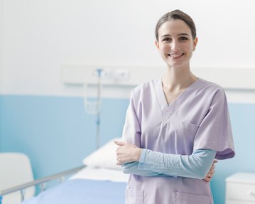 Eine Krankenschwester in lila Uniform steht lächelnd in einem Krankenhauszimmer neben einem Bett.